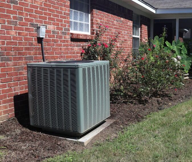 An AC unit outside a red brick home next to plants on a sunny day.